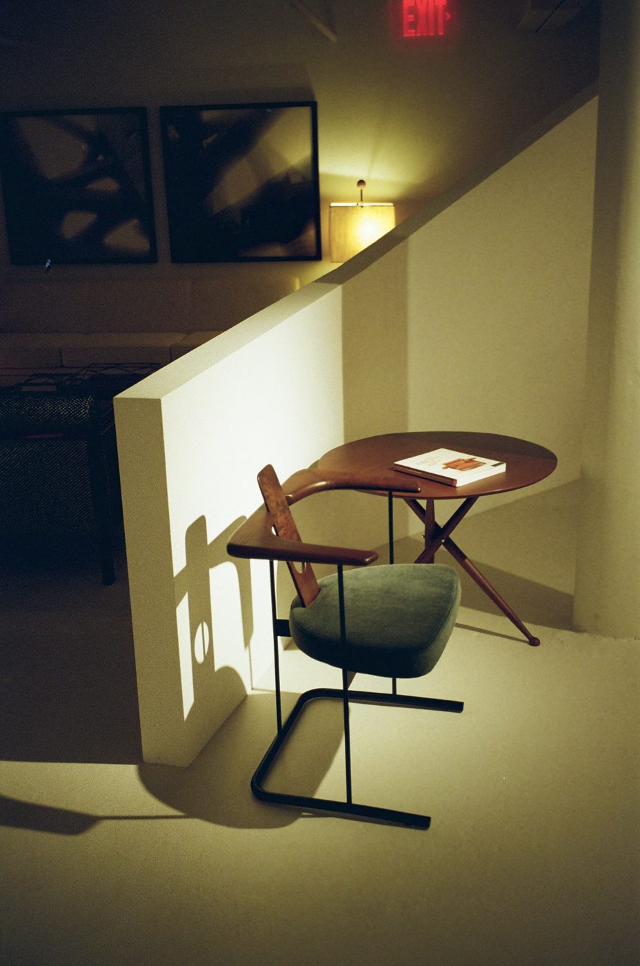 Wood and upholstered armchair beside a tripod side table in the Espasso gallery, lit under an exit sign