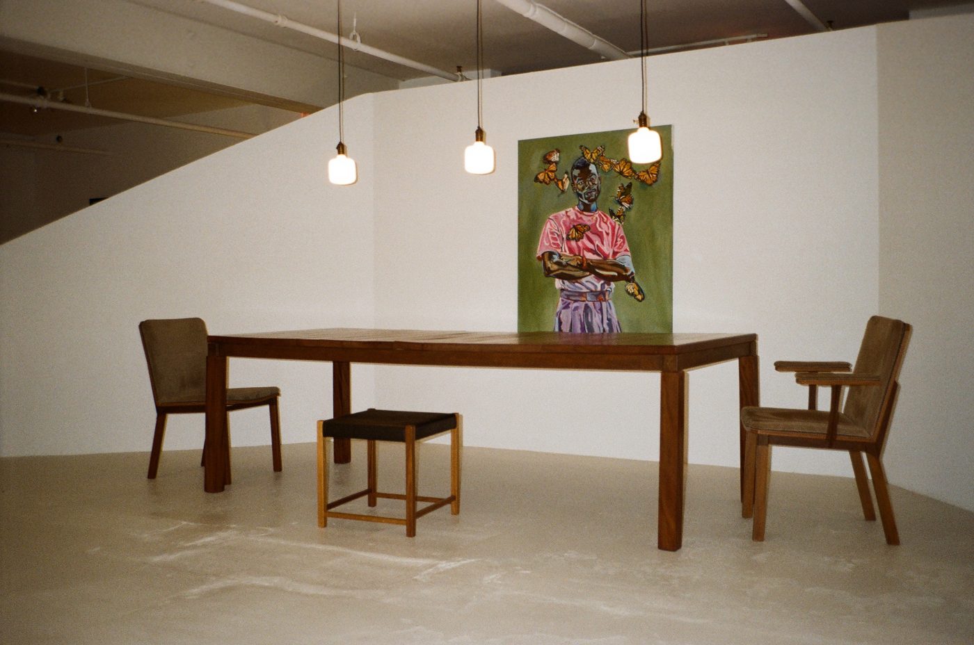 Solid wood dining table with chairs and a woven stool beneath pendant lights, a painted portrait on the wall