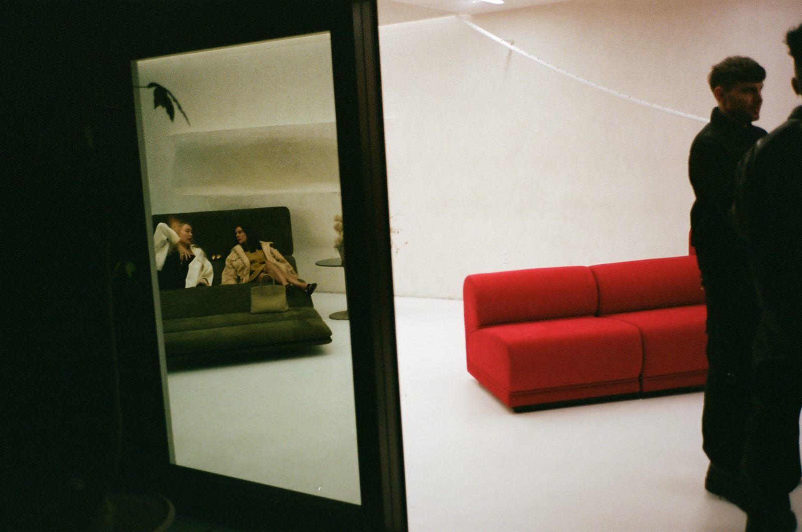 Gallery doorway reflecting two figures, a red modular sofa in the white room beyond