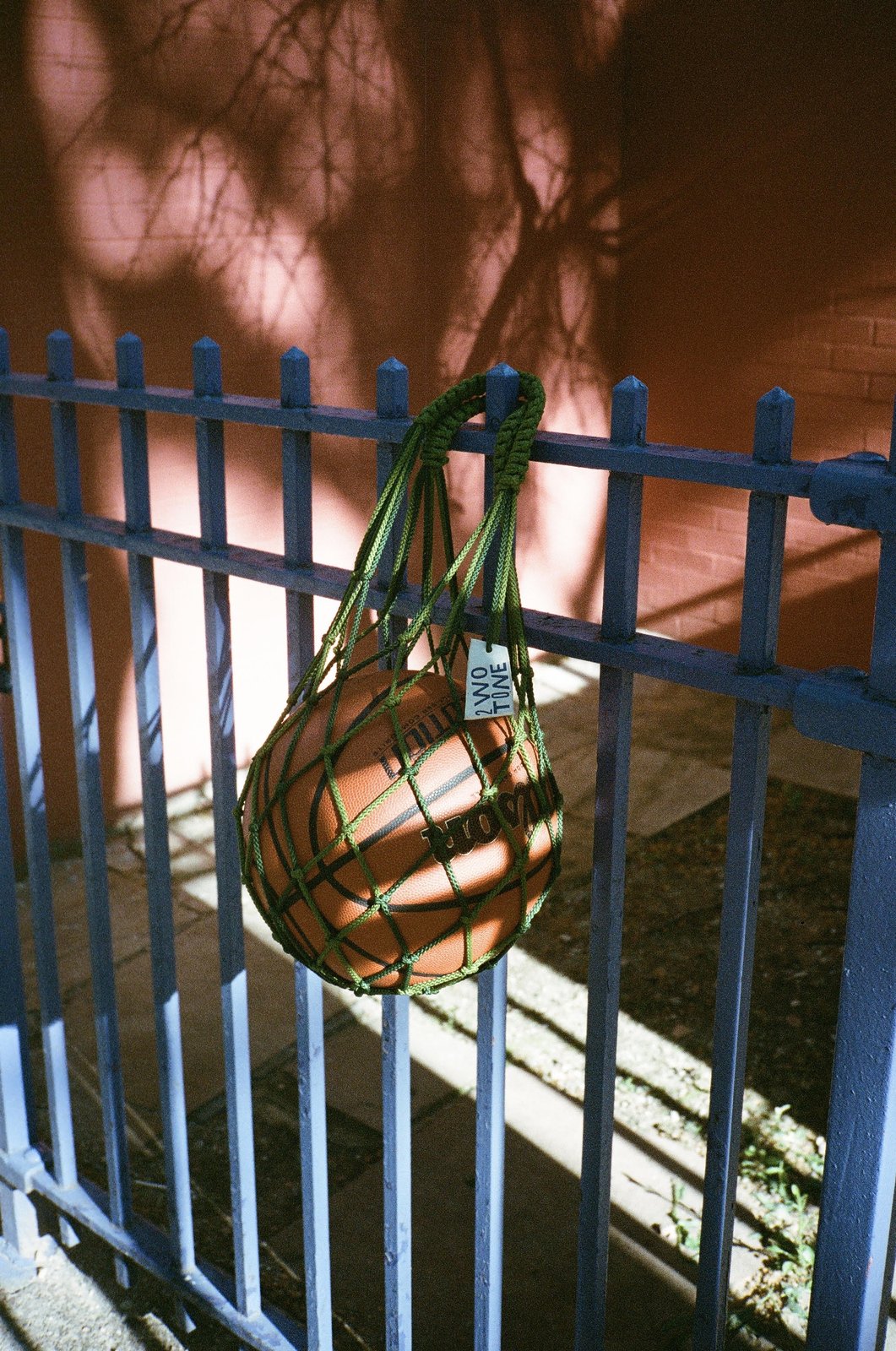 Basketball in a green rope 2WO TONE bag hanging on a blue iron fence, tree shadows on a red brick wall